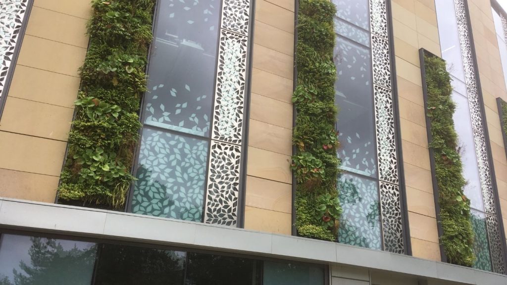 A modern building facade features vertical green walls with lush plants between beige panels and decorative metallic window accents.