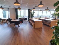 Modern office lounge with wooden floors, round tables and black chairs, large windows letting in natural light, indoor plants, and hanging pendant lights.