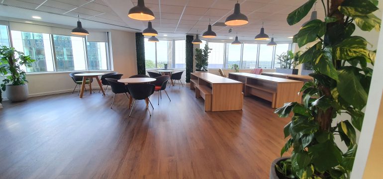 Modern office lounge with wooden floors, round tables and black chairs, large windows letting in natural light, indoor plants, and hanging pendant lights.