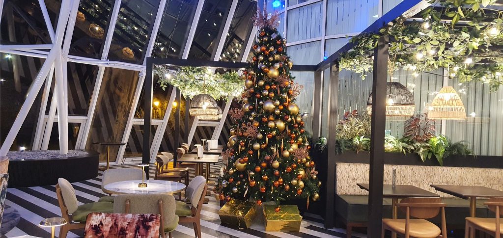 Large tall decorated Christmas tree inside a modern restaurant with windows, lamps and indoor greenery.