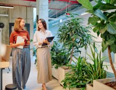 Two women with tablets walk and talk together through an office filled with potted plants.