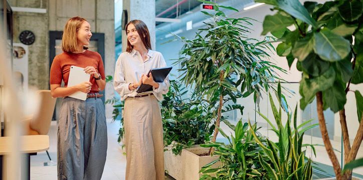 Two women with tablets walk and talk together through an office filled with potted plants.