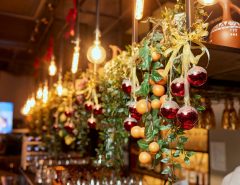 Christmas decorations including red ornaments, faux greenery, oranges, and gold bows hanging from a metal shelf above a bar.