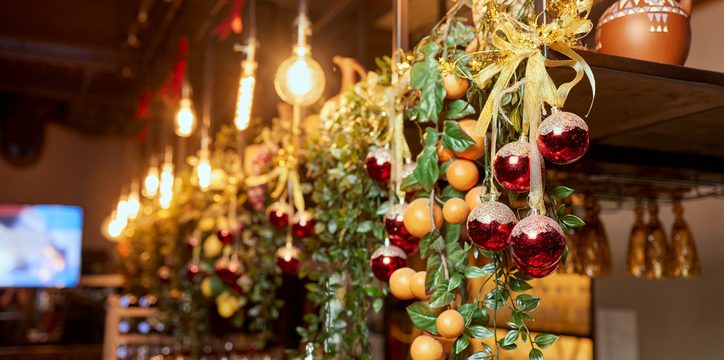 Christmas decorations including red ornaments, faux greenery, oranges, and gold bows hanging from a metal shelf above a bar.