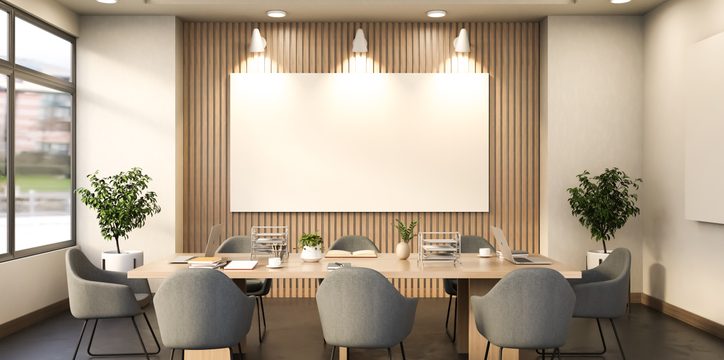 A conference room with a large empty rectangular frame mounted on a wood-paneled wall above a long table surrounded by six grey chairs.