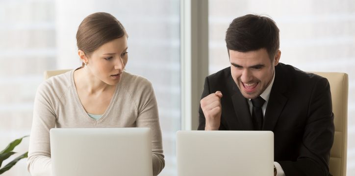 A woman looking surprised at her laptop while a man in a suit clenches his fist in celebration at his own laptop.